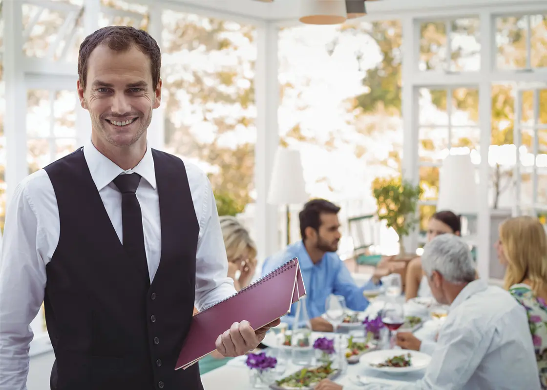Restaurantmeister mit Speiskarte vor einer gedeckten Tafel mit Gästen.